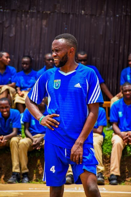 A male athlete in a blue soccer uniform on a sunny sports field with spectators in the background.